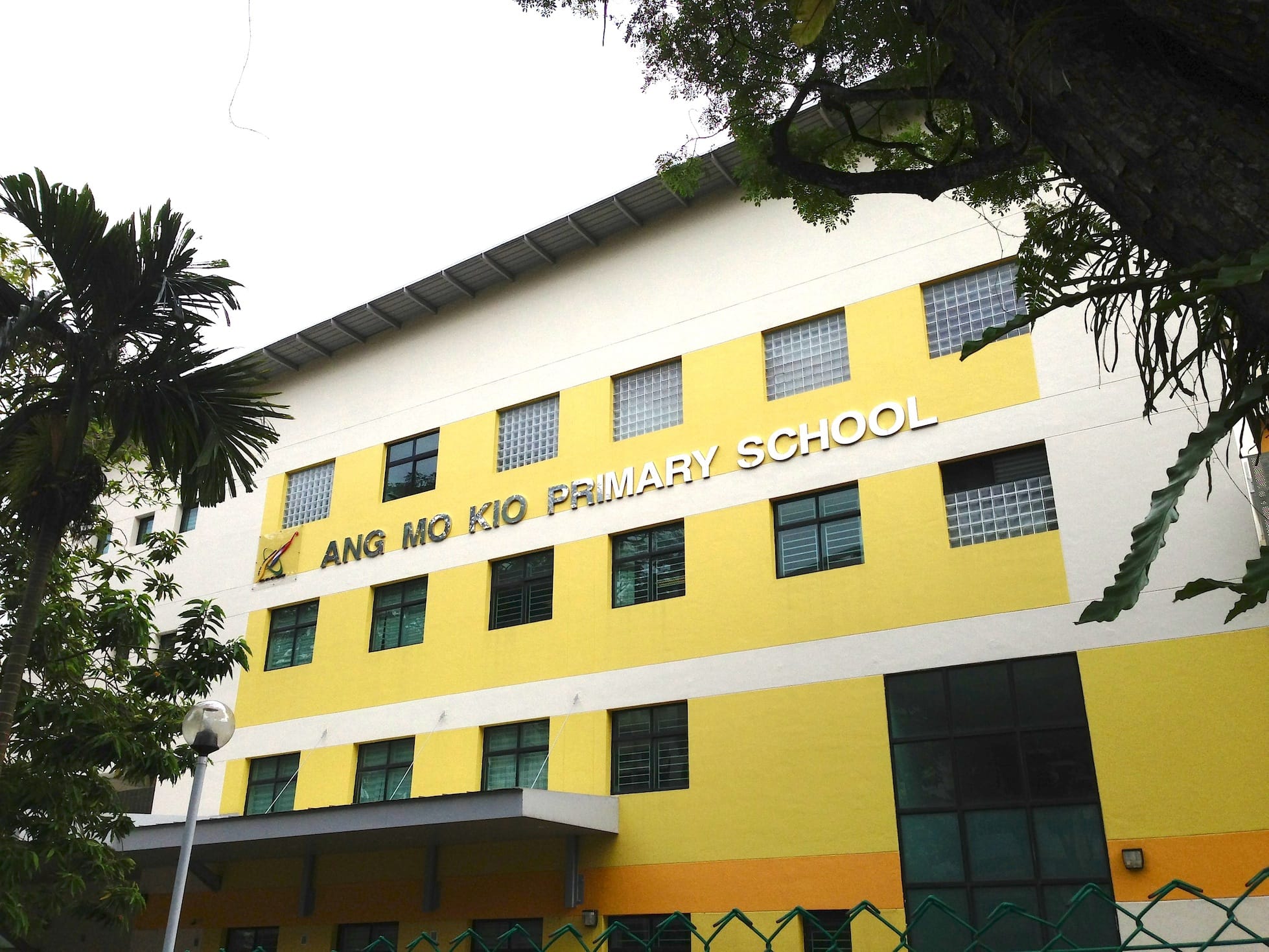 Yellow and white multi-storey building with "Ang Mo Kio Primary School" sign, surrounded by trees.